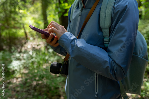 Close-up of female traveler using smartphone in the forest with camera and backpack. Navigation, hiking, travel app and outdoor lifestyle concept
