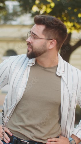 Serious dissatisfied Caucasian man tourist in glasses standing outdoors looking for someone waiting for meeting angry frustrated looks time, touching wristwatch feeling upset about being late lateness