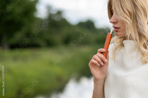 Young woman enjoying a vape outdoors by a river on a sunny day with green trees in the background
