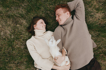 A couple lies on a grassy field, smiling while holding a small white goat kid between them. Cozy sweaters, natural light, and relaxed mood convey a lifestyle moment of farm life, bonding, and gentle