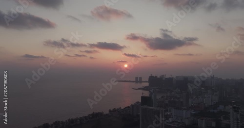 Backward aerial view: the sun sets over the calm sea, casting a golden glow across the Sihanoukville coastline and the hazy city skyline.