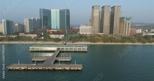Panning aerial view of a coastal pier with a helipad topped glass terminal extending into the sea, set against the Sihanoukville skyline of high-rise hotels and apartments.
