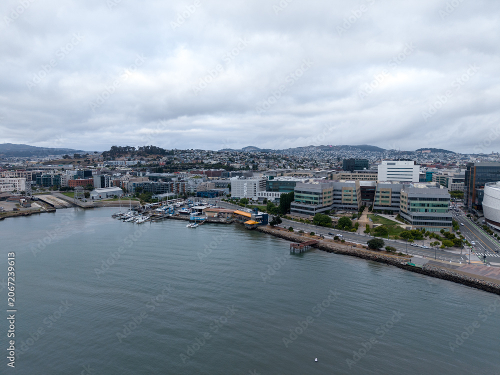 Obraz premium Drone view capturing the Mission Bay neighborhood along the waterfront with modern buildings and a marina foregrounding distant hills, showcasing urban development and coastal living