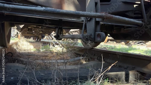 Low view beneath a freight railway wagon showing rusted steel wheels, bogie and suspension above an overgrown disused track. Gritty industrial rail-freight infrastructure
