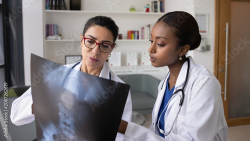 Younger African American doctor asking senior trauma surgeon for advice, teamwork, showing Xray of spine, pointing at image. Two medical professionals discussing scanning exam results. Banner shot