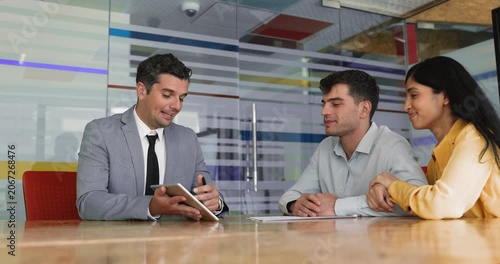 Happy diverse husband and wife visiting lawyer, real estate agent, financial and tax advisor. Friendly male professional showing content on tablet computer to couple of clients