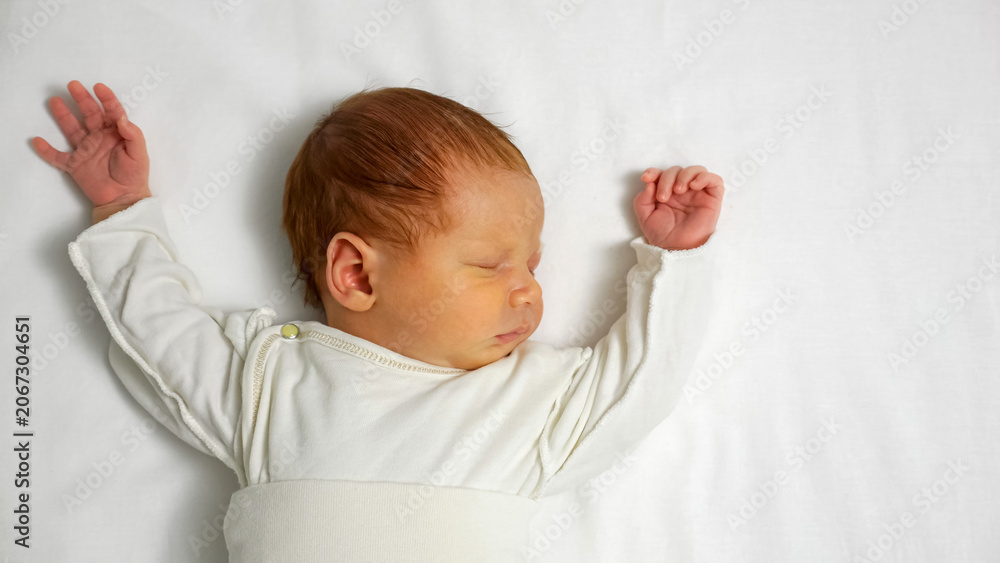 Obraz premium Newborn baby with red hair sleeping profoundly in comfortable white clothes and stretching arms on a clean white sheet, symbolizing innocence, new life, and peaceful dreams