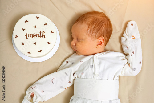 Newborn baby boy sleeping peacefully next to a small white cake decorated with 1 month text and tiny hearts, celebrating growth and early childhood
