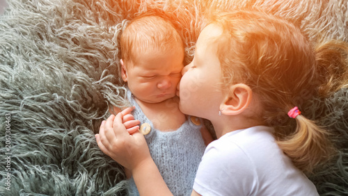 Young girl showing affection and love to her newborn brother or sister, holding hands while lying together on a soft furry blanket with warm sunlight
