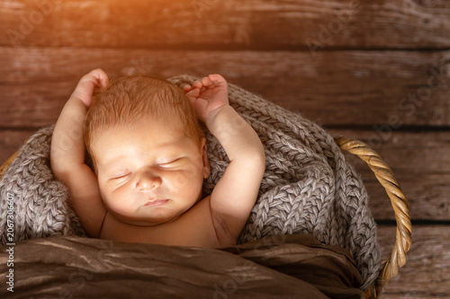Newborn baby boy sleeping calmly in a rustic woven basket covered by soft gray knit and brown fabric, representing innocence, new beginnings, safety, and a tender moment