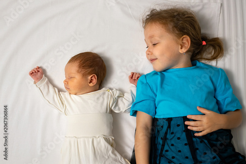Sleeping newborn baby lying next to her older sister on a white bed, symbolizing family love, childhood innocence, and the bonding between siblings