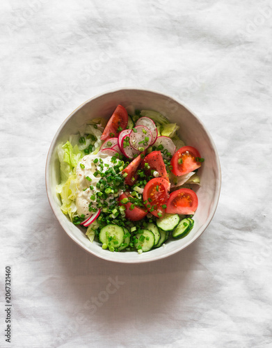 Fresh salad of garden vegetables - tomatoes, iceberg, cucumbers, radishes and yogurt dressing on a light background, top view