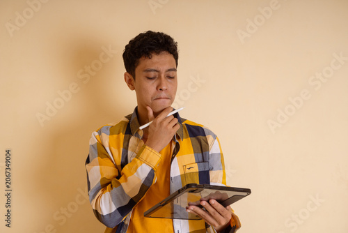 Contemplative Young Man in Checkered Shirt Holding Tablet and Pondering Thoughts
