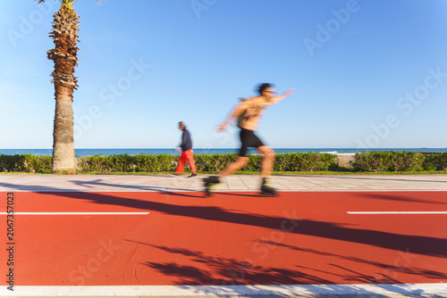Fotografija Man rollerblading quickly along a red asphalt path next to the ocean on a clear