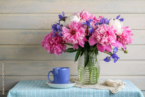 Rustic flower still life with pink peonies and irises in glass vase, cup and lace on table. Cozy summer home decor with soft floral mood.