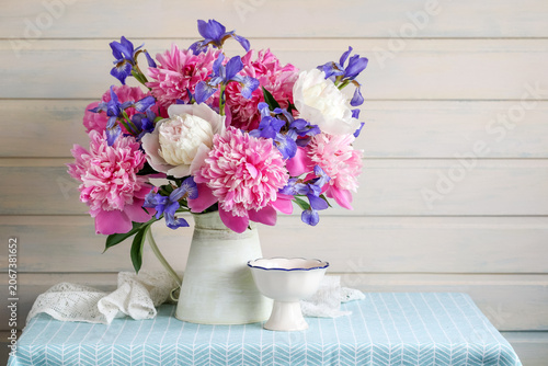 Light rustic still life with pink peonies, purple irises in tin pitcher, white bowl and lace on table, on pale wooden background.