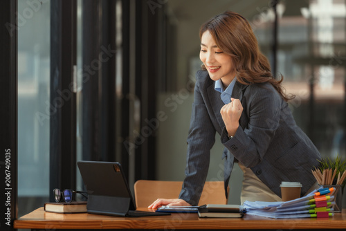 Cheerful Asian businesswoman celebrating success with fist pump looking at tablet in office.