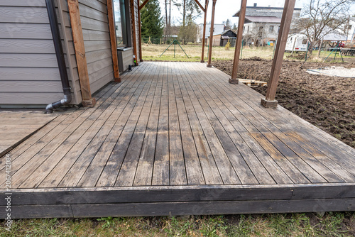 A weathered wooden deck shows sun bleached boards, dark stains, diagonal boards meeting straight runs, simple posts, drainage piping, bare trees, and cool diffuse daylight.