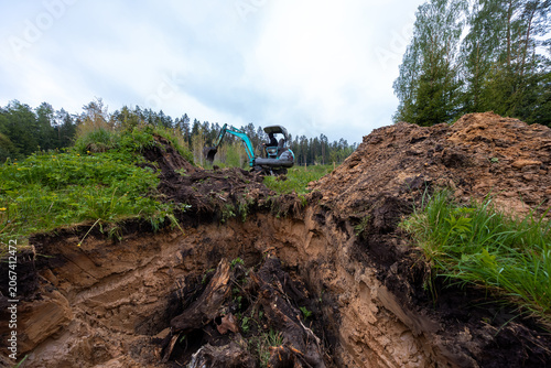A teal excavator works on an open dig site in a boreal forest clearing, exposing stratified sand, peat, and fresh root mass under overcast daylight from a low wide view.