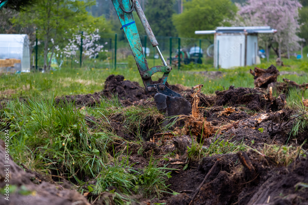 Fototapeta premium A mini excavator arm digs a rough trench, prying roots and stumps. Wire fencing, a white greenhouse tunnel, and a shed with solar panels sit under soft overcast light.
