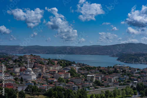 A panoramic view of a residential neighborhood overlooking the Bosphorus, featuring colorful houses, a mosque, green hills, and a dramatic sky filled with clouds. The landscape highlights the unique b