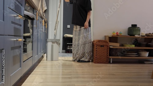 A teenager handles a mop in the kitchen while doing household chores on a light wooden floor.