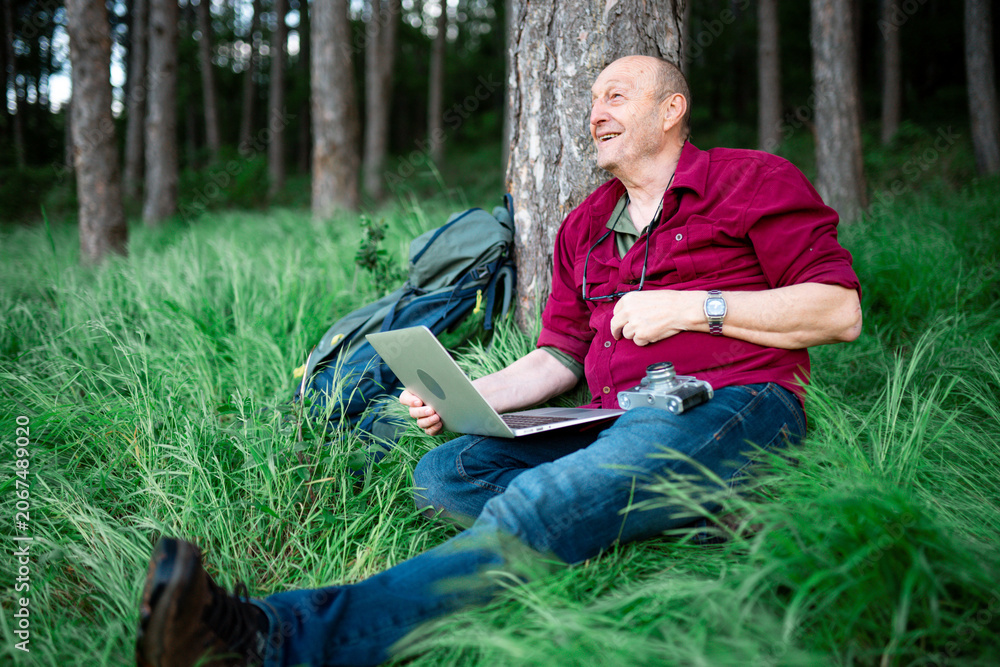 Obraz premium An elderly man with a laptop and a camera sits on the grass leaning against a tree and excitedly rejoices at the good news about the photos he sent online