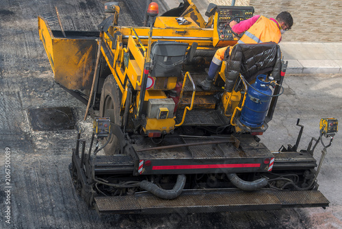 High angle view of an asphalt paver machine laying hot black tar on a city street, operated by a road construction worker in a safety vest.