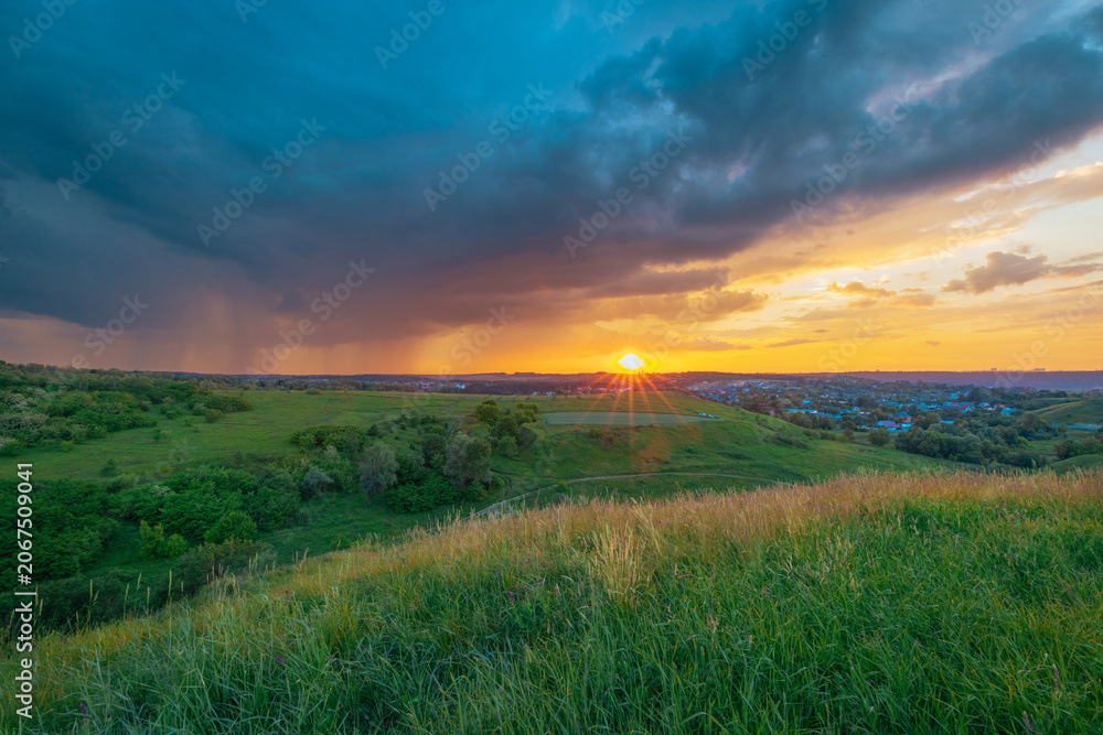 Fototapeta premium Stormy clouds over the horizon. Summer sunset over the green grass field. Approaching rain in the evening. 