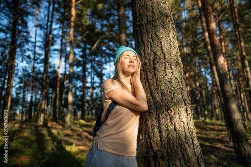 Fotografie Relaxed woman touching tree trunk in autumn forest, closed eyes and basking at sunlight