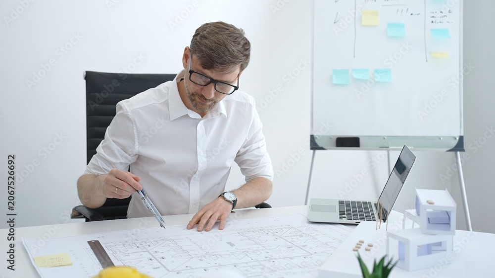 Naklejka premium Male architect drawing on building blueprints with a compass, developing architectural plans for a new house model, and working on construction design in a modern office