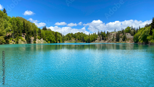 Wide panoramic view of Green Lake in Georgia with turquoise waters, dense evergreen forest and bright summer sky. Calm reflections and untouched wilderness create a serene mountain landscape.