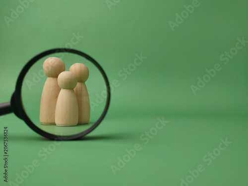 Three wooden peg figures standing together on a green background are focused through a black magnifying glass frame