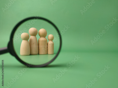A close-up view of a hand holding a magnifying glass over three wooden peg figures on a green surface