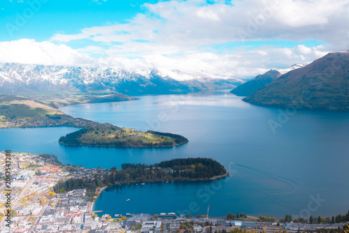 Queenstown, New Zealand: Wide horizontal landscape travel panorama over Lake Wakatipu, green peninsula and city below snow-capped alpine mountains.