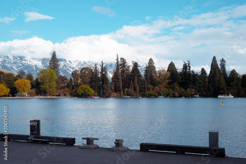 Queenstown, New Zealand: Concrete wharf pier edge with travel view of glittering blue Lake Wakatipu and dense green autumn forest trees under clouds.