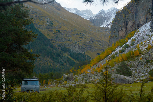 Grey off-road vehicle parked in a mountain valley with rocky slopes and snow-capped peaks. Autumn trees with yellow foliage, rocky terrain. Wild nature, overland travel, and adventure atmosphere. 