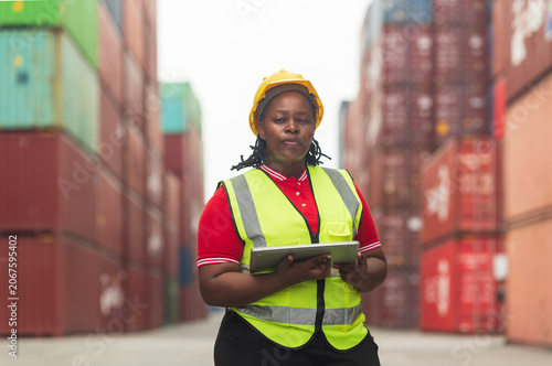 Black foreman woman worker talk on walkie-talkie at container yard, holding laptop computer. Logistics import export shipping concept.