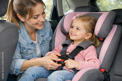 A mother sitting next to her young child strapped into a booster seat inside a car. They are both smiling.