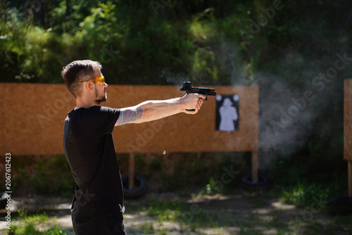 Focused young man practicing target shooting with black pistol outdoors. Firearm safety training, shooting range marksmanship drill, tactical defense posture, active sport shooting discipline