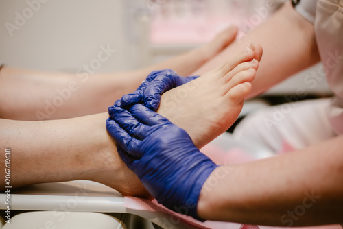 Feet care, a pedicurist wearing blue gloves for hygiene is performing foot care on a female client inside a feet spa clinic.