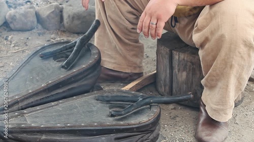 Historical reenactment of an artisan in traditional clothing using antique leather bellows to fan the flames in an ancient outdoor workshop.