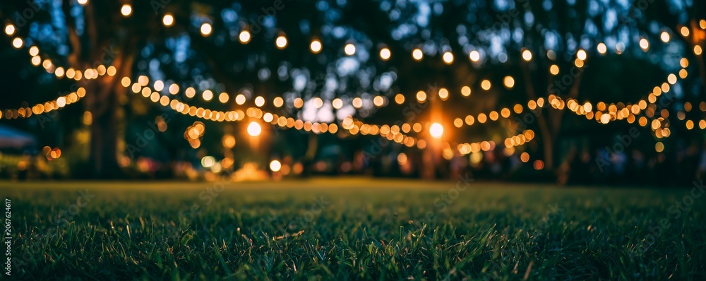 Fototapeta premium Close-up of fresh green grass with bokeh string lights glowing warmly in blurred background at an outdoor evening event, concept for festive celebrations, wedding decor and garden parties