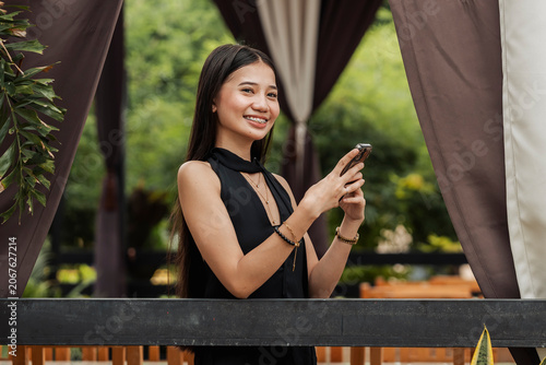 Canvastavla A young Southeast Asian woman smiles confidently at the camera while holding her smartphone in an outdoor alfresco setting
