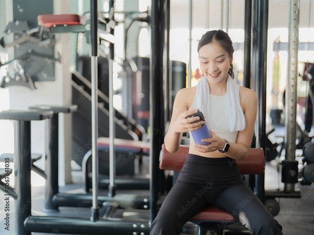 Fototapeta premium Smiling young Asian woman in sportswear holding a water bottle and sitting on a bench press in the gym after a workout.