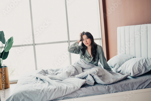 Φωτογραφία Tired young woman sitting up in bed holding her head, waking in bright bedroom l