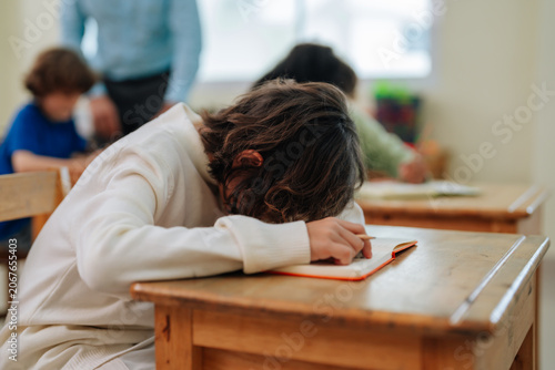 Student boy sleeping head down on desk during school lesson with teacher standing background inside classroom environment