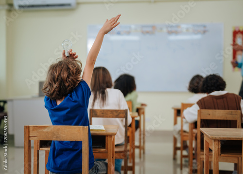 Student boy raising hand and holding lightbulb above head during classroom lesson with teacher standing at whiteboard background