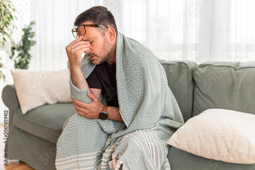 Man feeling unwell and exhausted, experiencing a persistent headache or migraine while resting on a comfortable seating, covered with a patterned throw, needing medical care