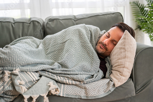 Man sleeping under a warm blanket, finding comfort and peaceful repose on a fabric couch, indicating relaxation and needing recovery